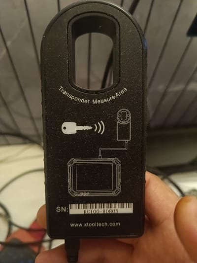 A person's hand is holding a black electronic device with white text and graphics. The device has a U-shaped handle at the top. The text on the device reads "Transponder Measure Area", "SN: KC100-80805", and "www.xtooltech.com". A graphic depicts a key with radio waves emanating from it, pointing towards a rectangular object with circular elements, which is then connected by lines to a larger rectangular outline. A barcode is visible next to the serial number. The background appears to be a blurry indoor setting with vertical lines, possibly a grid or a slatted surface. There are also some cables visible in the lower left corner of the image. The image is taken from a close-up perspective, focusing on the device and the hand holding it. No specific location in Huntingdonshire, United Kingdom, can be identified from the image.