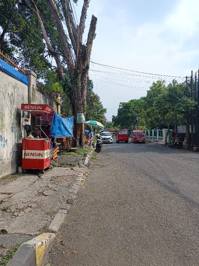 This image depicts a street scene in Bojongherang, Indonesia, observed from a low angle looking down the asphalt road. On the left, a damaged concrete sidewalk runs parallel to a concrete wall marked with graffiti. Two red mobile fuel vending stations, each displaying "BENSIN" (gasoline), are positioned on the sidewalk; one features a pump mechanism, the other is a simpler box. Behind these, a blue tarp covers a makeshift stall, and a large, heavily pruned tree trunk rises, supporting multiple overhead utility wires. Further along the sidewalk, a green market umbrella indicates another informal vendor setup. A black scooter is parked near these stalls.
The asphalt road, appearing two-laned, is visible leading into the distance. Several vehicles are present on the road: a white passenger car is partially visible on the left side, followed by two red passenger micro-vans, consistent with local public transportation, facing away from the viewer. On the far right side of the road, another scooter with a rider is partially visible near a dark, vertical slatted fence. The environment includes abundant green trees and foliage lining both sides of the street, with a light blue, slightly overcast sky overhead.