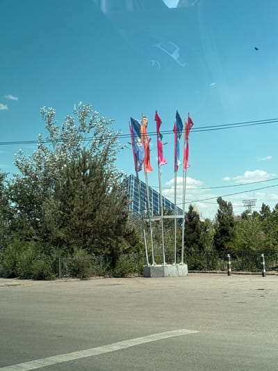 A bright, sunny day is depicted in this photograph, taken in Jezqazğan, Kazakhstan. In the foreground, a paved road with a white lane marker runs horizontally across the frame. To the left of the road is a patch of dry grass and a low metal fence. Beyond the fence, a group of trees are visible, with a prominent pine tree in the center and a tree with lighter colored leaves to its left. In the middle ground, a cluster of colorful flags on poles stand tall. Behind the flags, a modern building with a glass facade and a distinctive angular roofline is partially visible. The background consists of a clear blue sky with a few wispy clouds. A solitary bird is in flight in the upper right portion of the sky. Reflections of a car's interior are visible on the surface of the sky, indicating the photograph was likely taken from inside a vehicle.