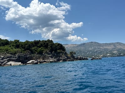 The image captures a coastal view with a body of blue water in the foreground. A rocky shoreline with dense green vegetation and trees lines the left side of the frame. In the background, there are mountains with scattered buildings and a clear blue sky with white clouds. The scene appears to be a natural landscape possibly located in the Kassopaia Municipal Unit, Greece, known for its scenic coastlines and mountainous terrain. There are no people or animals visible in the image. The overall impression is one of a serene and picturesque natural environment.