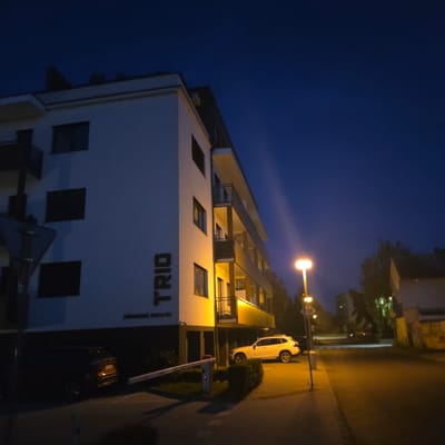 An exterior, low-angle, full shot captures a modern apartment building at night in Piešťany, Slovakia. The building, illuminated by a streetlamp on the right, features a white facade with multiple levels of balconies. The word "TRIO" is emblazoned vertically on the left side of the building. Below the building, a white automatic barrier arm is extended across a paved area. Two cars are visible: a dark-colored SUV parked to the left of the barrier, and a white SUV parked to the right, partially obscured by a hedge. The sky is a deep twilight blue. The scene is devoid of people.