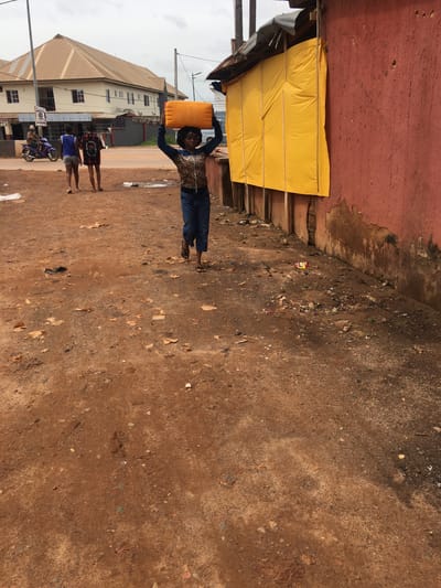 In Enyibichiri, Nigeria, a woman stands in a dirt street with a yellow rectangular container balanced on her head. She wears a leopard-print top and dark pants. Behind her, two children stand near a parked motorcycle. A multi-story building is visible in the background, while a yellow makeshift awning covers a section of a red wall to the woman’s left. The ground is uneven with scattered debris.