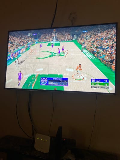 The image captures a television screen displaying a basketball video game, likely set indoors in a dimly lit room. The game features the Sacramento Kings playing against the Milwaukee Bucks.

On the top right of the screen, the scoreboard indicates "SAC" (Sacramento) has 13 points and "MIL" (Milwaukee) has 10 points. The game is in the "1st" quarter with "2:05" minutes remaining, and a number "23" is also visible, possibly indicating a shot clock. A digital player, identified as "G. Antetokounmpo" (Giannis Antetokounmpo), is visible on the court, highlighted by a ring around him. On the bottom left of the screen, player statistics for the "SACRAMENTO KINGS" are shown: "D. FOX" has 9 "PTS" and 4/4 "FES", while the "REST OF TEAM" has 4 "PTS" and 2/5 "FES". The virtual basketball court features the Milwaukee Bucks' branding, including "FEAR THE DEER".

To the right of the television, a white internet router or modem with several green indicator lights is visible. Various black cables and wires run behind the TV and connect to the router and other dark electronic devices situated below it. The ambient lighting of the room is dim, suggesting it might be evening or a low-light environment. No specific visual cues within the image confirm the provided location context of Lakka, Sierra Leone.