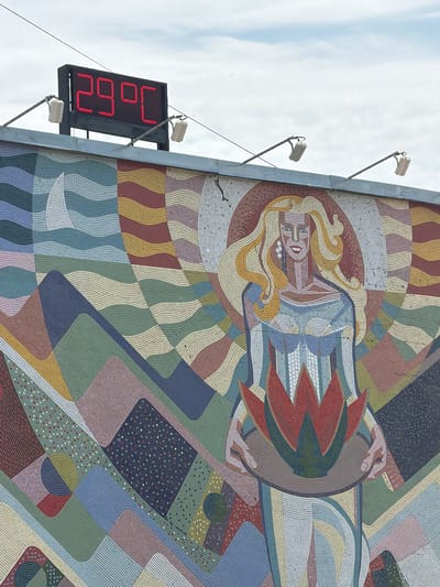A medium shot captures a segment of a mosaic mural, seemingly affixed to a building wall in Kamyshin, Russia. A red LED display adjacent to the mural shows "26.9°C". The mosaic features a woman with blond hair, enclosed in a circular design. A sun-like element with red and green rays surrounds her. The mosaic's background consists of geometric shapes in various colors, including red, yellow, green, and blue. The sky is visible above the mural, indicating an outdoor setting. The image appears to be taken during daylight.