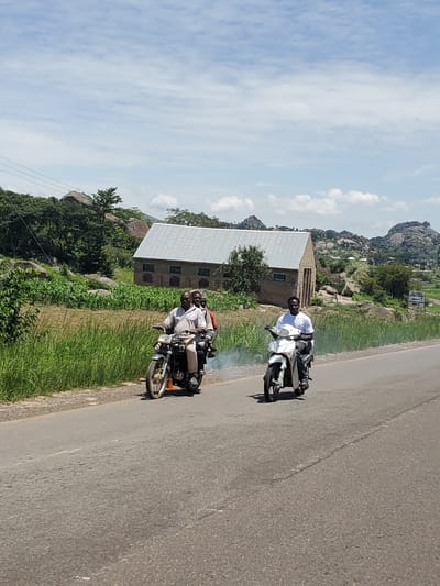 In Pankshin, Nigeria, two motorcycles are traveling on a paved road. On the left, a motorcycle carries three men; the driver is wearing a light-colored collared shirt and light pants, and is looking towards the camera. The two passengers behind him are partially obscured. On the right, a man wearing a white t-shirt and dark pants is driving a white scooter, also looking towards the camera. Both vehicles appear to be in motion, with smoke emanating from the exhaust of the motorcycle on the left. The road is bordered by a grassy verge and a field of crops. In the background, a building with a corrugated metal roof stands among trees and large rock formations, with a mountainous landscape visible beyond. The sky is partly cloudy.