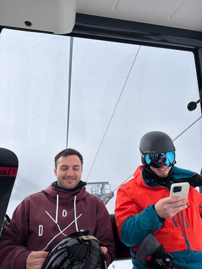 Two adult males are observed inside a ski gondola or cable car, which is in motion through an overcast and wet environment at a ski resort near Andermatt, Switzerland. The windows are covered in numerous raindrops, indicating precipitation.
The male on the left, with dark hair and a slight beard, wears a maroon hooded sweatshirt featuring a white "DOD" logo and geometric design. He holds a black, wet ski helmet in his lap with both hands, looking downwards with eyes partially closed and a slight smile. A black snowboard with red accents and "Head" branding is propped vertically beside him to his left.
The male on the right wears a black ski helmet with "sandbox" branding and blue mirrored goggles, largely obscuring his face. He is dressed in an orange ski jacket with teal/blue accents and black ski pants. He looks down at a white smartphone held in both hands, with a visible smile beneath his goggles.
Outside the gondola, a thick steel cable runs vertically. A metal ski lift tower structure is partially visible through the rain-streaked windows and cloudy, foggy background, consistent with a mountainous ski area.