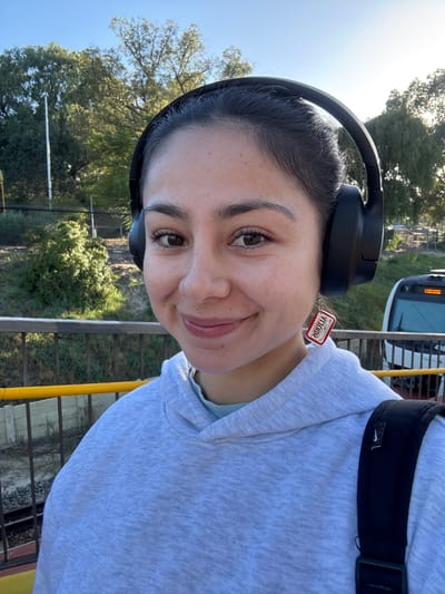 A person stands on a platform, likely at a train station in Bassendean, Australia. They are wearing black headphones, a light grey hooded sweatshirt, and a black backpack with a visible Nike logo. A red tag with reversed writing hangs off the headphones, near the person's left ear. The person is looking towards the camera with a slight smile. The environment includes a train in the background, trees, a fence, and yellow railing. The location is consistent with a train station in Bassendean, Australia, considering the type of train and surrounding vegetation.