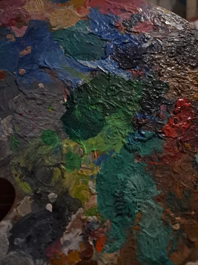 The image displays a close-up view of a painter's palette, heavily laden with various colors of oil paint. The paint is thickly applied, creating a textured, almost sculpted surface. Dominant colors include deep greens, blues, reds, and browns, interspersed with lighter shades of grey, beige, and yellow. The paint is mixed and swirled, showing the history of recent artistic activity. The texture suggests the paint is still wet or has recently dried, with visible ridges and peaks formed by the brushstrokes. There are no people, scenery, or specific events depicted, only the abstract arrangement of colors on the palette. The lighting appears to be artificial, casting subtle shadows that accentuate the paint's texture. The overall impression is one of creative potential and the raw materials of art. No text is visible in the image. The location context of Popovo, Bulgaria, provides a geographical reference but is not visually evident in the image itself.
