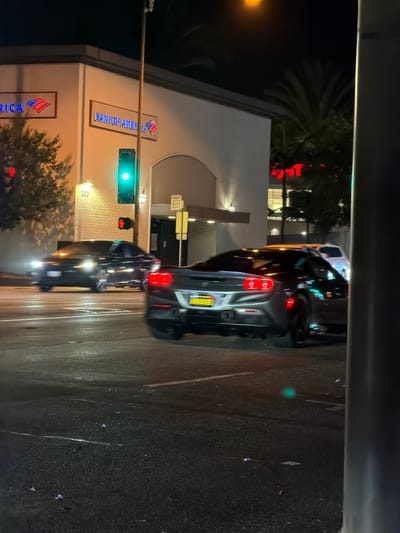 At night, on a wet street in Glendale, California, a dark sedan drives through an intersection. The traffic light is green. The sedan's brake lights are illuminated. In the background, a Bank of America building is visible. A palm tree stands behind the bank. The street is illuminated by the glow of streetlights and car headlights.