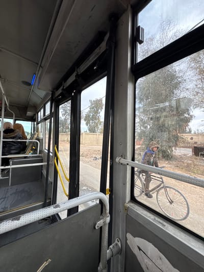 The media file presents a view from inside a public transport vehicle, looking out through its upper windows. The interior of the vehicle features metal framing, overhead bars, and a small blue light fixture on the ceiling. Yellow hanging straps or handles are also visible inside.
Through the multiple window panes, an outdoor scene is observed under an overcast sky. The ground outside appears to be unpaved, possibly dirt or gravel, with scattered trees showing bare branches, indicating a cooler season like late autumn or winter. In the mid-ground, distant structures or buildings can be seen.
The primary subject visible outside is a person riding a bicycle. The individual is dressed in camouflage-patterned clothing, including a jacket and a patterned hood or head covering. A blue strap or harness is worn across their chest. The bicycle is a standard type, with visible spoked wheels. The person appears to be actively riding the bicycle on a path parallel to the vehicle.
Inside the bus, in the lower portion of the image, the back of a person's head and shoulder are partially visible. They are wearing a jacket with a fur-lined hood. Next to them is a black trash bag, possibly containing personal belongings.
The scene captures an ordinary moment of transit in Ashrafiyat Sahnaya, Syria, with a passerby on a bicycle outside. The weather is cloudy, and the overall impression suggests a non-urban or semi-rural setting. There is no visible text in the image.