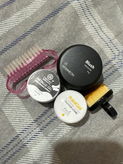 The image displays a close-up, overhead view of several nail art products arranged on a grey, checkered fabric. The objects include two nail brushes, one pink and translucent with a curved handle, and another with a black handle and yellow bristles. There are also three small containers of nail gel. One is a black container labeled "WOWMEOW Blush UV/LED GEL 15g". Another is a white container with a black lid, labeled "CRISMO PROFESSIONAL METALLIC GEL ЗЕРКАЛЬНЫЙ ЭФФЕКТ SILVER 5gr". The third is a white container with yellow lettering that reads "PaintGel Золотой оскар 5ml". The background fabric is a soft grey material with a pattern of alternating thick white and thin blue stripes running diagonally across it. The lighting is even, suggesting an indoor setting. No people are visible in the image. The location in the city of Яромінскі сельскі Савет, Belarus is not discernible from the visual information provided.