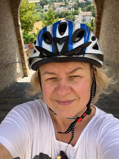 A woman wearing a white t-shirt and a blue and white bicycle helmet is taking a selfie on a stone staircase. The woman has blonde hair and is smiling. The background shows a view of houses and trees in the city of Barbara, Italy. The staircase is made of stone and has a metal railing on the left side. The woman is wearing a gold necklace.