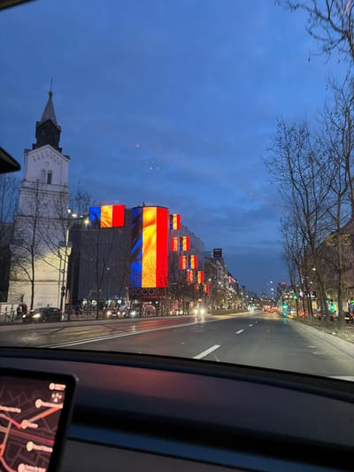 The image depicts an urban street scene at dusk or early night in Jilava, Romania, viewed from inside a vehicle. In the immediate foreground, part of a vehicle's dashboard is visible, featuring a digital screen displaying a red-orange map interface.
The main subject is a multi-lane asphalt road, appearing wet and reflecting artificial lights, with white lane markings stretching into the distance. On the left side of the road, a tall, white building with a steeple, characteristic of a church or clock tower, stands prominently. Adjacent to it is a modern commercial building with several large, illuminated LED panels integrated into its facade, displaying the vertical blue, yellow, and red stripes of the Romanian flag. Text reading "CCCOR" and "WRLD.WIDE.ALL" is visible on the lower section of this building. Pedestrians are present on the sidewalk in front of these structures, and several cars are visible on side streets and the main road.
On the right side of the street, a row of bare deciduous trees with visible branches lines the sidewalk. Beyond the trees, commercial buildings with illuminated storefronts and signs extend along the road. Streetlights are active, casting light on the environment. Moderate traffic is observed on the road, with headlights from oncoming vehicles and taillights from receding ones. The sky overhead is a deep blue, indicative of twilight or early evening, with scattered cloud formations.