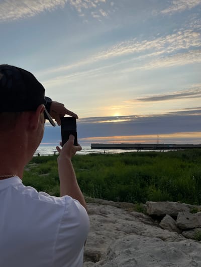 A person, viewed from behind, stands on a rocky embankment and takes a photo of a sunset over the sea with their smartphone. The person wears a black baseball cap, a white t-shirt, and a gold chain. The sun is low in the sky, peeking through scattered clouds, casting a warm glow over the water. A long pier extends into the sea, with a few people visible on it. Green grass grows on the embankment in the foreground, with large rocks scattered around. The sky is a mix of blue and orange hues, with wispy clouds. The scene appears to be a serene, coastal landscape.