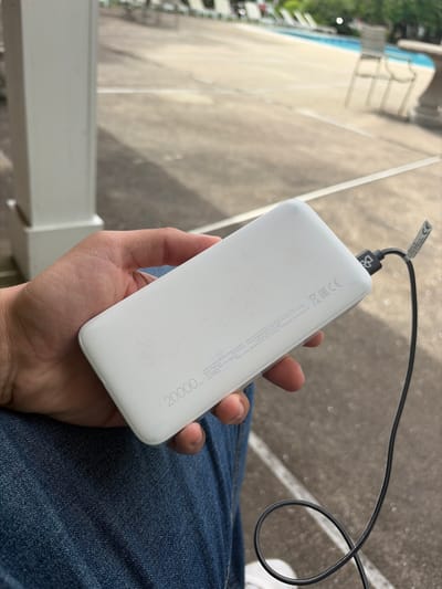 A white power bank, labeled "20000 mAh," is held in a hand over a pair of blue jeans. A black charging cable is plugged into the device. The background features a gray concrete area with architectural elements like a white railing. It is impossible to determine if the image was taken in Edison, United States of America.