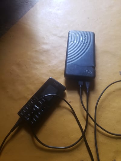A black feature phone is lying on its side on a wooden surface and is plugged into a dark blue power bank with a black charging cable. The power bank has a circular design on its surface and the brand name "itel" is visible. The phone displays the brand name "LAUN" and has a keypad with numbers and symbols. The wooden surface appears to be a table or floor with some dust particles. The background is partially visible and shows a dark, possibly shadowed area. The scene is located in Rigasa, Nigeria.