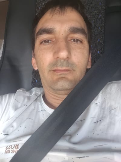 A male subject is depicted in a close-up, head-and-upper-torso shot, seated within the interior of a vehicle. The specific location in the city of Velleminfroy, France, cannot be determined from the visual information provided.

The subject is an adult male with short, dark brown hair and an olive skin tone. He has brown eyes, a straight nose, thin lips, and exhibits light stubble on his chin and upper lip. His eyebrows are dark and somewhat prominent. He maintains a neutral facial expression, looking directly forward into the camera.

He is wearing a white short-sleeved t-shirt adorned with a repeating pattern of small, dark, short horizontal lines and dots. Partially visible text, appearing inverted and possibly reversed, is printed on the lower left portion of his shirt, reading "ANEO BOSS 220R" or similar characters. A black, ribbed seatbelt crosses his chest diagonally from his upper right shoulder towards his lower left side.

Behind the subject, a vehicle seat is visible. The left side of the seat appears to be dark grey or black, possibly leather or synthetic material, while the right side shows a dark upholstered fabric with a pattern of small blue or light-colored square or dot elements. The presence of the seatbelt and the vehicle-like seating indicates the subject is inside a form of transportation, such as a car or bus. The lighting is even, suggesting an enclosed or diffused environment.