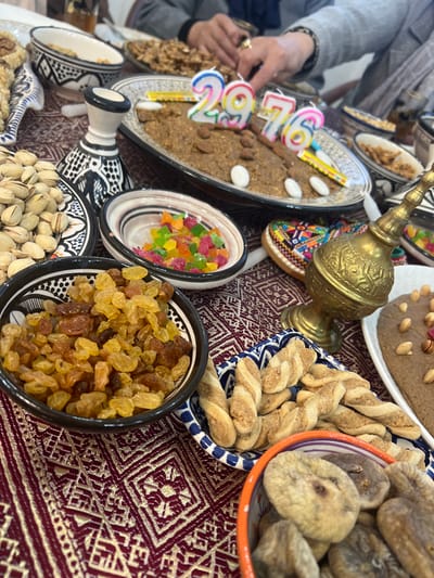 A table displays various foods including a light brown cake with birthday candles spelling out "29", along with almonds and white candy. Surrounding it are bowls of dried figs, raisin, candies, and pistachios. A silver pot sits beside these, while a dark brown cake with almonds decorates another section. Twisted pastries are also visible, and the table is covered with a maroon and white patterned cloth.