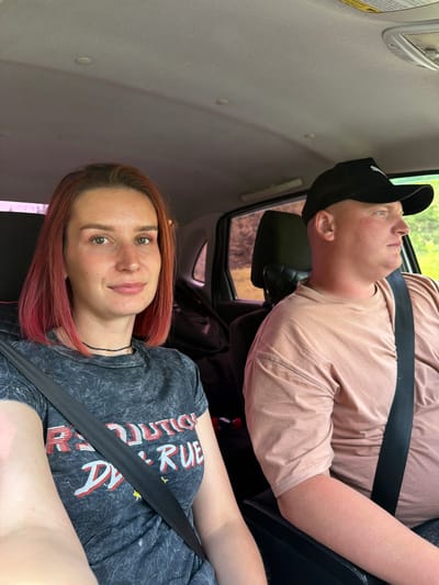 Two people are in a vehicle. The person in the front passenger seat is looking at the camera. This person has reddish-brown hair and is wearing a gray t-shirt with a design. The person in the back is looking up and toward the driver's side window. This person is wearing a black baseball cap and a pink t-shirt. Both are wearing seatbelts. The interior of the vehicle is light gray. The location is possibly the city of Горняк, Russia, but this cannot be confirmed from the image alone. The environment appears to be daytime.