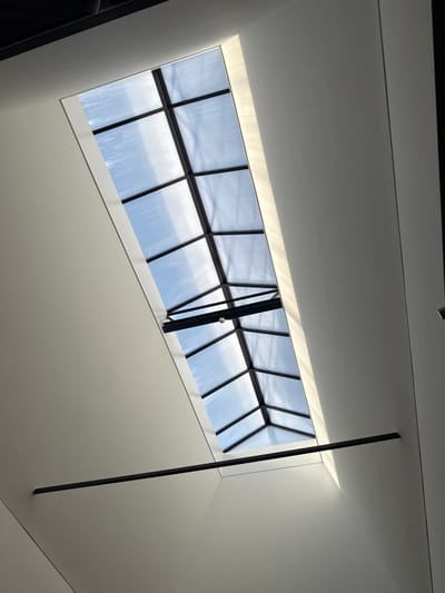 The image presents an upward view within a modern interior located in Harderwijk, Netherlands. The central subject is an elongated, rectangular skylight set into a light-colored (white or off-white) ceiling structure. The skylight features a dark, likely black metal frame creating a grid pattern, supporting translucent, corrugated glazing. Through this glazing, a clear blue sky with minimal cloud cover is visible. The skylight appears to be a pitched or gabled design, with a small, white cylindrical light fixture mounted on a horizontal cross-beam within its framework. Natural sunlight is entering through the skylight, casting distinct, parallel linear patterns of light onto the interior wall surfaces, particularly prominent on the right side of the frame. Below the skylight, a single black horizontal bar extends across the width of the ceiling space, perpendicular to the skylight's orientation, attached to opposing walls. No individuals or other discernible actions are present within the frame. The immediate environment consists of clean, unadorned architectural surfaces.