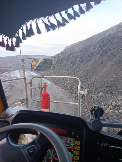 The image, taken from the perspective of someone inside a vehicle, captures a view of a quarry. A black fabric with dangling tassels is visible at the top of the frame. In the foreground, a steering wheel and part of a dashboard with various indicators are present. Outside the vehicle, a dirt road winds through the quarry. A red fire extinguisher is mounted on a railing to the right of the road. In the distance, large piles of dark-colored rock and excavated earth form the quarry walls. The sky is overcast, with a bright horizon. There are no people visible in the immediate foreground. The location is identified as Kyrgai, Russia.