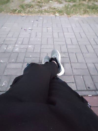 The image shows a close-up view of a person's legs and feet, from the knees down, with their legs crossed. They are wearing dark pants and grey sneakers with white soles. The person is sitting on a paved area made of grey rectangular stones, which appears to be a sidewalk or pathway. To the left of the paved area, there is a strip of grass with some fallen leaves, suggesting an outdoor urban environment. The lighting is dim, indicating it might be dusk or an overcast day. The image has a grainy quality. There is no information in the image to confirm the location as Stryków, Poland.