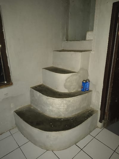 The scene depicts an interior view of a building located in Tarqumiyah, Palestinian Territory. A set of L-shaped stairs, constructed from concrete or masonry, dominates the central frame. The lower three steps feature prominent rounded, outward-curving risers, with dark-colored, worn treads. The upper steps become more rectilinear and recede into a corner. Two blue, cylindrical beverage cans are placed on the third step from the bottom. The surrounding walls are light-colored, likely plastered, exhibiting some discoloration and marks. A dark-framed window is partially visible on the left side of the image, and a dark wooden door frame is present on the right. The floor in the foreground consists of white square tiles with dark grout lines. No individuals are present in the image.