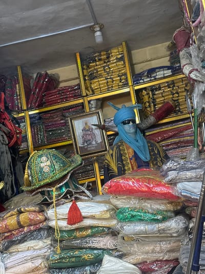 The image displays the interior of a market stall in Tudun Wazurchi, Nigeria, specializing in traditional attire and textiles. The environment features light-colored, untextured walls and ceiling, with a single exposed LED bulb providing illumination.

Gold-colored shelving units line the rear of the stall, densely packed with stacks of individually plastic-wrapped textiles in various colors, including red, yellow, and blue patterned designs. A light-skinned mannequin, dressed in a light blue turban-like headdress, dark sunglasses, and a dark tunic with prominent gold and blue embroidery, is positioned centrally on the right side of the frame. Behind the mannequin, a dark shaft with a red-wrapped tip, possibly a traditional spear or staff, is visible.

To the left of the mannequin, a framed portrait of a formally dressed figure, identified by the text "ALH. AMINU HAMZA (Wal Akbari)", hangs on the wall. The foreground is dominated by extensive piles of folded and plastic-wrapped embroidered fabrics in shades of red, white, green, gold, and silver. Two ornate green and gold traditional headdresses or hats, one featuring a red tassel and an intertwined gold cord, are prominently displayed on top of the textile pile in the lower-left quadrant. No human subjects are present within the scene.
