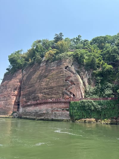 The image shows the Leshan Giant Buddha in Shizhong, Leshan, Sichuan, China. The colossal Buddha statue is carved into a cliff face adjacent to a river, likely a tributary of the Yangtze. A wooden staircase structure runs along the side of the cliff face, allowing visitors to ascend and descend. The cliff face is composed of red and brown rock formations, with vegetation, including trees, growing atop and along sections. Numerous people are seen on the staircase, suggesting the site is a tourist attraction. The sky is clear and blue. The river water is a greenish color.