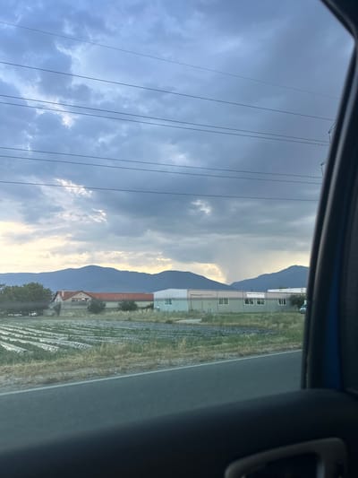 The image depicts a rural landscape viewed from inside a vehicle in Lozen, Bulgaria. A dark, cloudy sky dominates the upper portion of the frame, with several utility wires stretching horizontally across it. In the mid-ground, a field is laid out with rows of what appear to be covered crops. Behind the field, several buildings are visible, including a residential-style structure with a red tile roof and a longer, industrial-looking building with multiple windows. Further in the distance, a range of mountains forms the horizon. Notably, a visible rain shower can be seen falling from the clouds over the distant mountains. The foreground is filled with the interior of the car, including a portion of the door and a car handle, suggesting the photo was taken by a passenger.