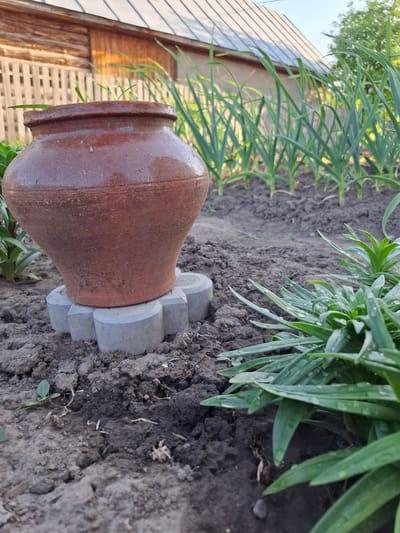 The image shows a brown clay pot resting on a concrete base with a flower-like design. The pot and base are situated on dark soil, likely in a garden or cultivated area. Green plants are growing near the pot. In the background, a wooden structure, possibly a fence or a shed, is partially visible. The location is specified as Амзя, Russia, but there are no visual cues in the image to confirm this location. There are no people visible in the scene. The subject is a decorative arrangement of a clay pot on a concrete base, situated in a garden or cultivated area. The focus is on the objects and their placement within the environment.