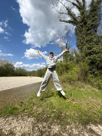 A person is captured in mid-air, likely jumping or posing dynamically, with their arms and legs outstretched in a star shape. The individual, wearing a grey hooded tracksuit, a light-colored baseball cap, and white sneakers, is smiling broadly at the camera, conveying a sense of joy and freedom.
The scene is an outdoor natural environment in Beregovoe, Russia, under a bright blue sky dotted with scattered white, fluffy clouds. The person stands on the edge of a wide, light-colored gravel bank, which runs alongside a clear, shallow body of water, resembling a river or canal. A strip of green grass borders the gravel bank on the left. Beyond the water and grass, the landscape is rich with lush green trees and bushes, indicating a vibrant natural setting. Some trees further in the background show bare branches, suggesting either early spring or a mix of tree types. The bright, sunny daylight indicates it is likely mid-day with pleasant weather. There is no visible text in the image.