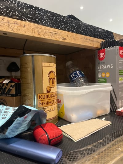 A cluttered storage space, possibly a drawer or cabinet, contains various items. A cylindrical tin of "Kurukahveci Mehmet Efendi Turkish Coffee" dominates the lower portion of the image. Beside it is a blue package with "UI COAREA" partially visible. A red, round object (perhaps a ball) and a slim, blue device are adjacent. Above the coffee tin is a clear plastic container, partly obscuring a "Ice Valley" water bottle. "Cater Pro Paper Straws" (black) are visible in their packaging. There is a piece of cardboard among the items. The cabinet or drawer is constructed of light wood, and the lining is a black material with white speckles. The location is not discernible from the image.