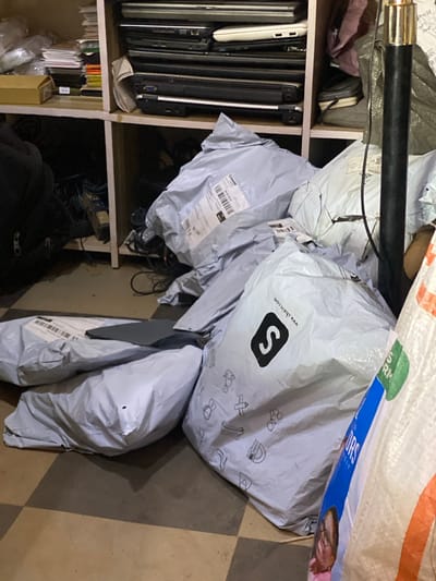 The image depicts a disorganized collection of packaged items and electronic devices, possibly in a storage area. In the foreground, several white and grey plastic mail bags are piled on a checkered floor. Some of these bags have visible labels, including one with the Shein logo and website. Behind the bags, stacked on shelves, are numerous laptops of varying colors, primarily black and silver. Some of these laptops appear to be older models. There are also stacks of paper and what look like electronic accessories or wires on the shelves. A black pole, possibly part of a lamp or stand, is visible on the right side of the image. The lighting suggests an indoor environment, possibly a storeroom or a retail back-office. The mention of Barakin, Nigeria, implies this scene is situated in that location.