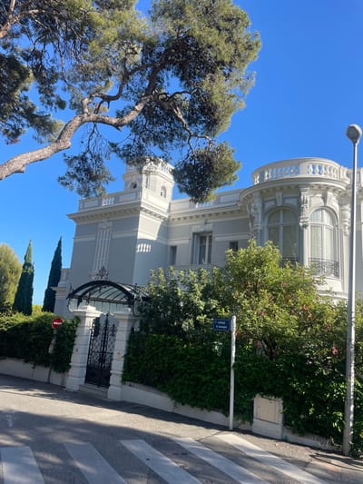 The image displays an exterior view of a multi-story villa located on Avenue R. Gramaglia in Cap-d'Ail, France, under a clear blue sky. The building features light gray walls with extensive white architectural detailing, including cornices, balustrades, and window frames. On the left, a square tower-like section has a small, arched window and a white balustrade at its summit. To the right, a prominent curved bay window section is topped by a white balustrade and intricate decorative elements. The main entrance, centered on the left side of the facade, consists of a black wrought-iron gate under an arched glass canopy with decorative ironwork, flanked by white stone pillars.

The property is bordered by a high, dense green hedge and various trees, including tall, slender cypress trees visible in the background on the left. A large, gnarled pine tree branch extends into the upper left of the frame, partially obscuring the sky and the building's roofline.

In the foreground, a paved asphalt road with white crosswalk stripes slopes upwards from right to left. A white street sign pole bearing the text "AVENUE R. Gramaglia" is situated on the right side, with a white utility box at its base. A street lamp on a pole stands on the far right. A red circular "No Entry" sign is mounted on a separate pole near the property's entrance gate. No individuals or active interactions are present in the scene.