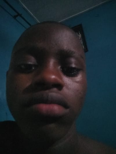 A close-up, low-angle portrait of a dark-skinned person with a shaved head. The person's face fills most of the frame. The person's expression is neutral. The background is a blurry blue wall. There is a framed object hanging on the wall. There is a black pole attached to the ceiling. It's impossible to confirm its location in the city of Epe, Nigeria.