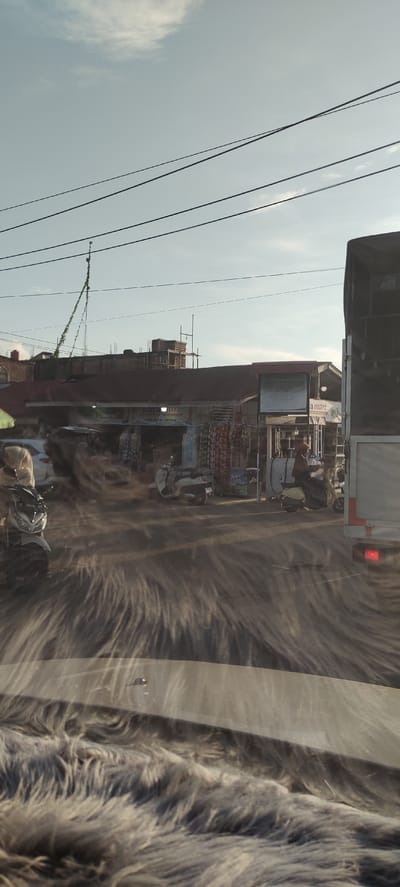 The image depicts a street scene in Meunasahmesjid, Indonesia. In the foreground, a car's windshield is covered with a fuzzy grey material, obscuring the view of the road ahead. The road surface itself is obscured by a dust or sand disturbance, creating a blurred effect that streaks horizontally across the image. On the left side of the road, a white motorcycle with license plate "BI 3655 GI" is parked. Further down the road, there are several shops with their fronts open to the street. One shop appears to be a "KARA MART". Scooters are parked in front of some of the shops, and people can be seen on them or walking. A large truck is parked on the right side of the road, its rear visible. Overhead, power lines crisscross the sky, and a vine-covered pole is visible. The sky is pale blue with some scattered clouds. The overall impression is of a busy street on a sunny day, with the dust disturbance adding a sense of movement or wind.
