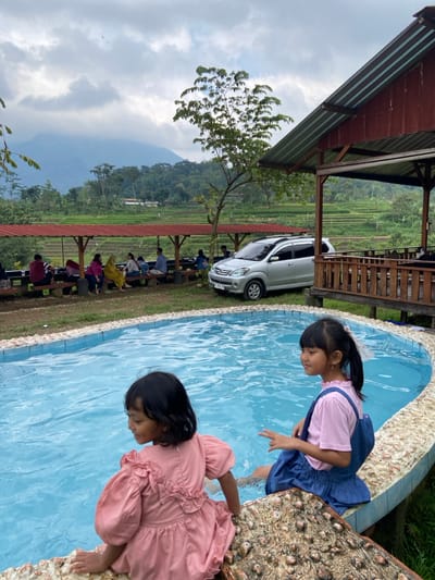 This image depicts an outdoor leisure area in Penanggungan, Indonesia, situated against a backdrop of terraced rice fields and lush mountains under an overcast sky. In the immediate foreground, two young girls are seated on the textured coping of a small, irregularly shaped swimming pool, their feet submerged in the clear blue water. The girl on the left, dressed in a pink ruffled dress, faces away from the camera, looking slightly left. The girl on the right, wearing a light pink shirt and a blue denim overall dress, faces right with her feet in the water.
In the midground, a silver Toyota Avanza is parked on a grassy patch. Beyond the vehicle, a series of wooden structures with red corrugated metal roofs house multiple individuals (approximately 10-12 discernible figures) seated on benches, indicating a social or dining setting. To the right, a larger, two-tiered wooden building with a darker roof is present. A tree with green foliage stands between the structures and the mountain range. The background reveals extensive green agricultural terraces, primarily rice paddies, ascending towards a prominent range of green-covered mountains. The sky is largely cloudy.