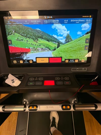 The image shows a treadmill with a large touchscreen display depicting a scenic view of a mountainside with a road and a truck. The treadmill console has various buttons for controls like "start," "program," "mode," and "stop." There are also controls for incline and speed. On the touchscreen, workout metrics are displayed, including "LUTNING" (incline), "HJÄRTFREKVENS" (heart rate), "KALORIER" (calories), "DISTANS" (distance), "STEG" (steps), "MINUTER" (minutes), and "HASTIGHET" (speed). The language on the display is Swedish. In the console's storage compartment, a small white object resembling a folded piece of paper or card is visible, along with what appears to be an Apple AirTag. The treadmill is situated indoors, on a dark floor surface with a wooden-patterned floor visible in the background. The image is taken from a slightly elevated perspective, looking down at the treadmill. The overall scene suggests a home gym or fitness center setting. There is no identifiable information that can place this scene in the city of Östersund, Sweden.