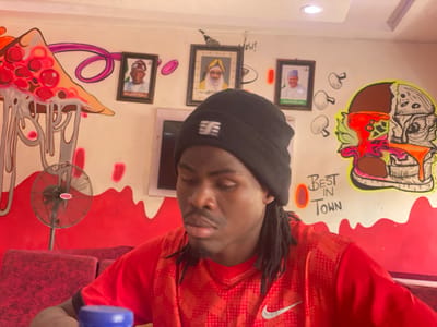 A man wearing a black beanie and a red shirt sits at a table in a restaurant in Tukur Tukur, Nigeria. Behind him is a wall decorated with food-themed graffiti, framed portraits of men, and a small television screen. An oscillating fan is also visible in the background. The restaurant wall is marked with the words "Best in Town".