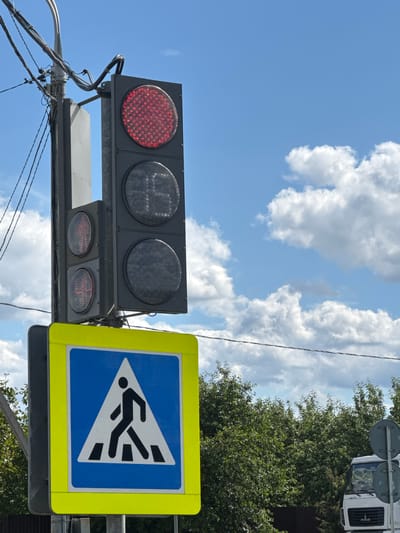 This media file shows a traffic light and a pedestrian crossing sign on a bright, sunny day. The traffic light has a red light illuminated, indicating that vehicles should stop. The pedestrian crossing sign is blue with a white triangle containing a black silhouette of a person walking across a crosswalk. The scene appears to be in an urban or suburban area, with trees and a portion of a white truck visible in the background. The sky is blue with scattered white clouds, suggesting good weather. The image is captured from a low angle, looking up towards the sky and the traffic control devices. There is no visible text in the media file. The overall impression is one of a typical street scene with traffic management in place.