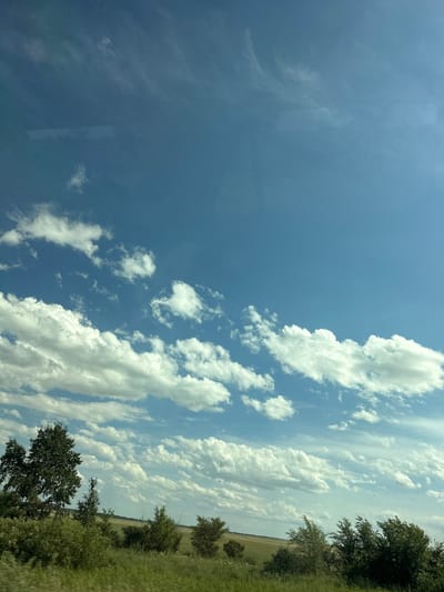 The image depicts a vast, clear blue sky with scattered white cumulus clouds. In the lower portion of the image, a landscape of green grass and trees is visible, suggesting an outdoor environment. The scene appears to be a natural landscape, possibly a rural area or a park. There are no people or objects visible in the image other than natural elements. The sky takes up the majority of the frame, with the landscape occupying the bottom quarter. The light suggests a daytime setting, possibly late morning or early afternoon, given the brightness of the sky and the illumination of the clouds. The overall impression is one of a serene and open natural vista.