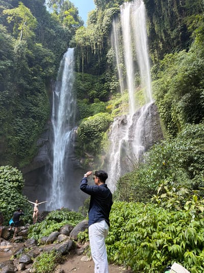 A man wearing a dark blue long-sleeved shirt and white pants stands with his back to the camera, holding his hand to his forehead as if shielding his eyes from the sun. He is positioned in the foreground, to the right of the center of the frame. Behind him, a wide, multi-tiered waterfall cascades down a lush, green cliff face. The water is misty and appears bright in the sunlight. In the middle ground, a woman in a bikini poses with her arms outstretched on a large rock in front of the waterfall. Another person, wearing a dark shirt and shorts, is partially visible to the left of the woman, also standing on rocks near the water. Several large rocks and dense green foliage surround the base of the waterfall and the individuals. The location is a natural outdoor environment, likely a jungle or tropical area, characterized by abundant vegetation and a prominent waterfall. The context suggests this scene is likely the Sekumpul Waterfall in the Sawan sub-district, Buleleng Regency, Bali, Indonesia, a popular tourist destination known for its impressive cascades.
