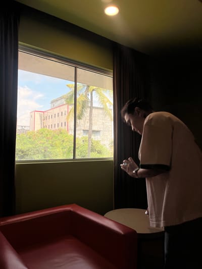 The image depicts an indoor scene looking out a window. A person, viewed from the side and partially in shadow, is standing and looking down at a mobile phone in their hands. They are wearing a light-colored t-shirt with dark trim on the sleeves. To the left of the frame, a dark curtain hangs, and in the foreground, a section of a red upholstered seating area is visible. Through the window, a building with a pale facade and red trim is seen, surrounded by lush green trees and palm trees. The sky above is a hazy blue with scattered clouds. The overall impression is of a room with a view of an urban or semi-urban environment. The lighting suggests it is daytime. The presence of palm trees and the architectural style of the building suggest a tropical or subtropical location, consistent with Dumai City, Indonesia.