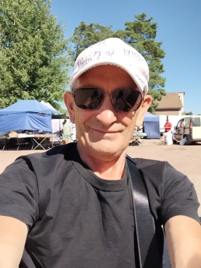 In a medium shot, a man wearing sunglasses and a white baseball cap with a patterned brim smiles at the camera. He is wearing a black t-shirt and a black strap is visible across his chest. The background depicts an outdoor market scene on a sunny day. Two blue tents are set up, with tables laden with merchandise. Further back, a white van and another tent are visible. The ground is a reddish-brown dirt or gravel surface. The man's head and shoulders are the primary focus, with the market serving as a backdrop. The location is not identifiable from the image alone as Irshansk, Ukraine.