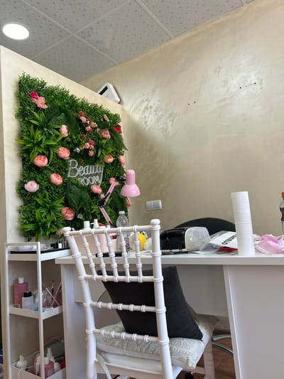 The image shows the interior of a beauty salon. A white desk is set up in front of a vertical garden wall featuring green foliage and pink flowers. A white chair with a dark cushion is pulled up to the desk. On the desk, there is a pink lamp, a roll of paper towels, several bottles of liquids, and a black device with a diamond-encrusted surface. To the left of the desk, a white shelving unit holds various beauty products. The walls are a neutral beige color, and the ceiling has a patterned tile design with a circular light fixture. A white electrical outlet and a small white device, possibly an air conditioner or purifier, are visible on the wall. The "Beauty Room" text is illuminated with neon lights on the vertical garden. The scene appears to be a well-organized and aesthetically pleasing workspace. The location is identified as Aksakovo, Bulgaria. There are no people visible in the image.