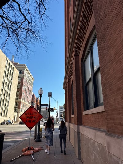 A street-level perspective shows two pedestrians walking on a sidewalk beside a brick building with large windows. Street signs and traffic lights are visible. A "Utility Work Ahead" sign is prominently displayed. Tall buildings are seen in the distance against a clear blue sky. A bare tree branch extends into the view on the left side of the frame.