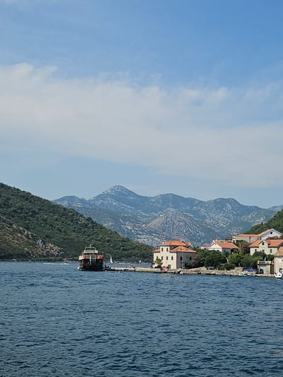 The image captures a serene daytime scene in Lepetane, Montenegro, featuring a waterfront view. In the foreground, the deep blue water of the bay is visible, with gentle ripples across its surface. On the left side of the image, a ferry is docked near a lush, green hillside. The ferry is dark-colored with a white upper deck and appears to be stationary.
In the mid-ground, a small coastal town with several buildings is situated along the water's edge. These buildings are characterized by their terracotta-tiled roofs and light-colored facades. Some of the buildings are residential homes, while others may be part of the town's infrastructure. A paved walkway or road runs along the waterfront in front of the buildings, with parked cars visible.
The background is dominated by a majestic mountain range, its peaks and slopes a mix of rocky grey and green vegetation. The sky above is a clear blue, with scattered white clouds adding depth and texture. The overall impression is one of a peaceful coastal settlement nestled between the mountains and the sea.