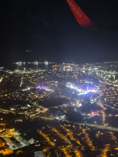 A nighttime aerial view from an EasyJet plane, with a city landscape. The city is illuminated by numerous lights, predominantly yellow and white. The angle indicates that the city is along a coast with water on the left side of the view. The city has many streets and buildings and a few areas of distinct purple and blue lighting. The image is blurry, indicating movement of the plane. The location is Matosinhos, Portugal.