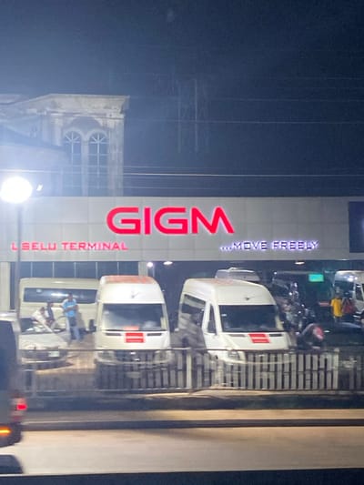 The GIGM (God is Good Motors) terminal is brightly lit. Red, vertical lettering spells out "GIGM L. SELLU TERMINAL …MOVE FREELY." Three white shuttle buses are parked at the terminal with passengers visible inside one. The image appears to be taken at night. A structure that looks like a church or a historical building is adjacent to the terminal. Streetlights create glare in the photograph. A fence is visible at the right.